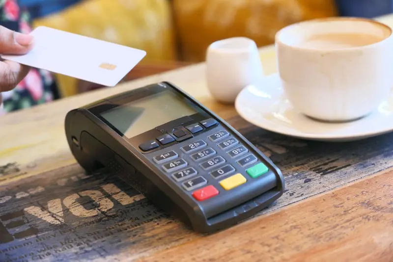 A hand holds a chip card over a countertop card terminal next to a cup of coffee