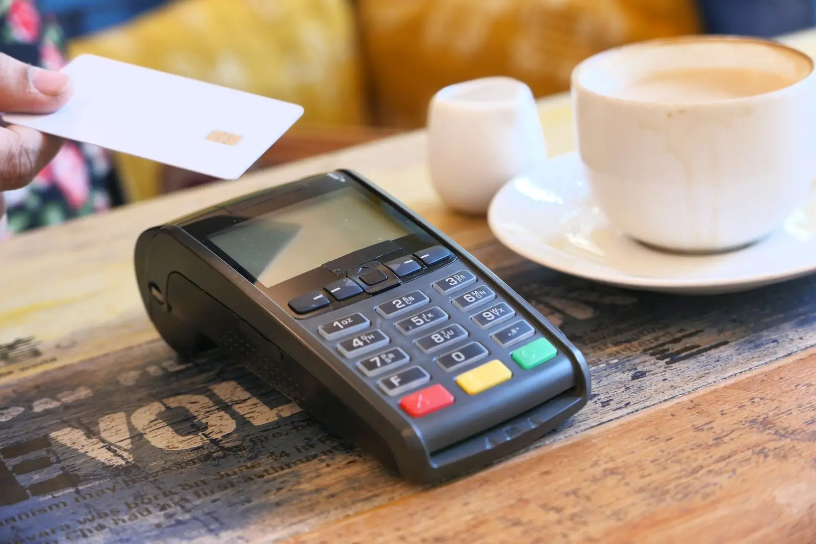 A hand holds a chip card above a countertop card terminal on a wooden cafe table, next to a cup of coffee