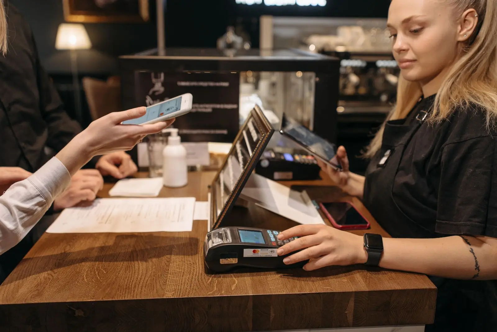 A customer holds a phone toward the card terminal on a wooden cafe counter while a staff member in a black apron enters the sale